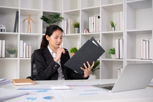 Focused Asian female accountant doing paperwork in office with plan documents on desk.