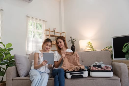 Young Asian woman packing clothes to the suitcase. Preparation for the summer vacation. Two women are planning and with using tablet for trip booking a trip and helping to prepare luggage to travel.