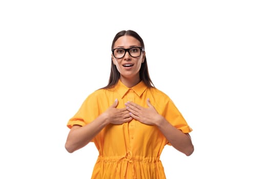 Caucasian young slender woman with black straight hair with glasses and in a yellow dress looks at the camera with surprise.