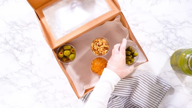 Flat lay. Hands are meticulously adding fresh red grapes to a bowl, complementing a beautifully arranged charcuterie box brimming with a variety of cheeses, olives, and cured meats, set against a sleek marble surface.