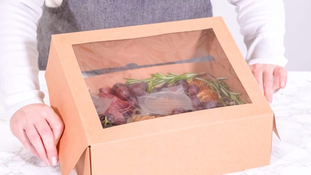 Hands are meticulously adding fresh red grapes to a bowl, complementing a beautifully arranged charcuterie box brimming with a variety of cheeses, olives, and cured meats, set against a sleek marble surface.