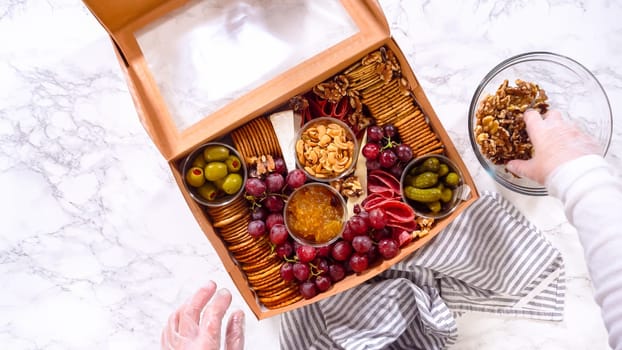 Flat lay. Hands are meticulously adding fresh red grapes to a bowl, complementing a beautifully arranged charcuterie box brimming with a variety of cheeses, olives, and cured meats, set against a sleek marble surface.