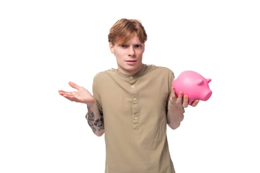 young red-haired man with glasses dressed in a brown shirt holds a pink piggy bank and decides where to spend his savings.