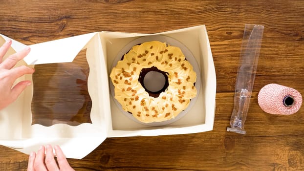 Flat lay. Carefully placing the gingerbread bundt cake, adorned with salted caramel frosting and gingerbread sprinkles, into a white paper bundt cake box with a clear window for gifting.