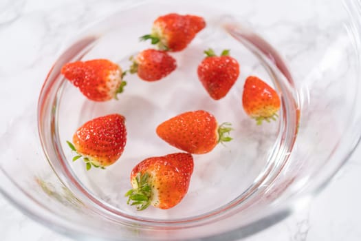 Ripe strawberries are submerged in water within a large glass mixing bowl, a step in washing the fruit to ensure cleanliness and longevity before storage or consumption.