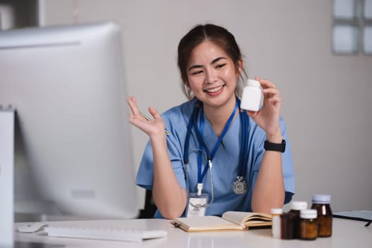 A woman in a blue scrubs is smiling and holding a pill bottle. She is sitting at a desk with a computer monitor in front of her