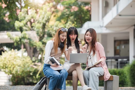 Group of happy young Asian college students sitting on a bench, looking at a laptop screen, discussing and brainstorming on their school project together..