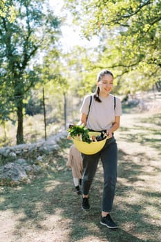 Little girl follows her mother carrying a bowl of greens and vegetables in her hands in a green park. High quality photo