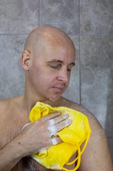 Bald man in the shower lathers his body with yellow washcloth in foam with cleanser