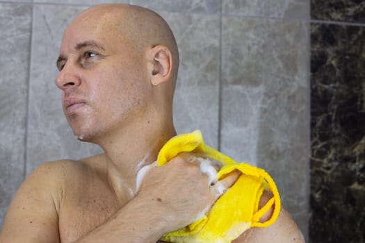 Adult bald man washes himself in shower with foamy washcloth, keeping men's body clean