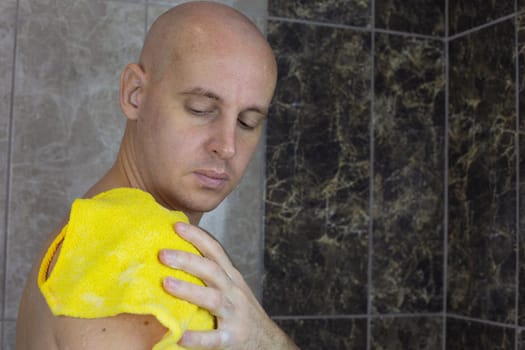 Man without hair washes himself in shower with washcloth, bald guy takes care of his skin in the shower with a special product with foam