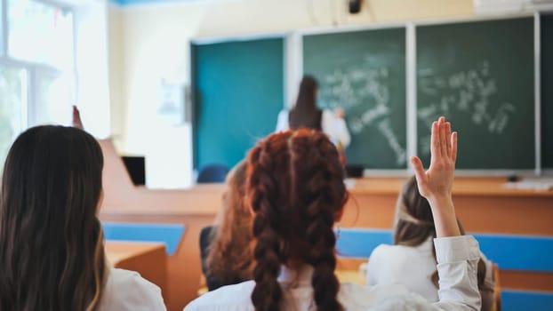 Intelligent group of young school children all raising their hands in the air to answer a question posed by the female teacher, view from behind