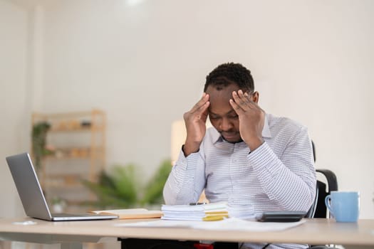 African American businessman feeling stressed while working on laptop, taking notes on documents in office and preparing work report..