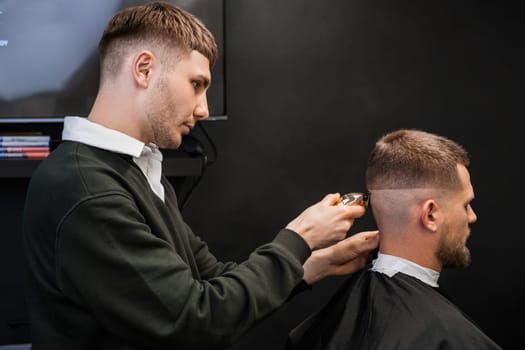 Young barber cuts mans hair using trimmer in the barbershop.