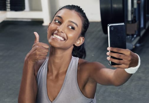 Woman, thumbs up and selfie on phone in gym for social media post or wellness blog in training. Happy, active and female person with mobile for sign of success workout or fitness session for health.