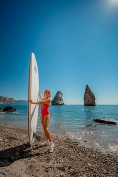 Woman surfer during summer travel to beach vacation. A middle-aged woman in a red swimsuit and a cap with sap on the background of the sea and rocks in the water. extreme sport. Travel, weekend, lifestyle
