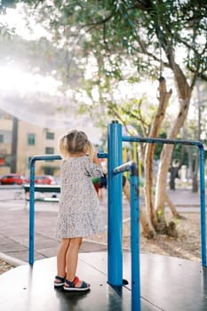 Little girl stands on the carousel holding the handrail and looks at the house. Back view. High quality photo