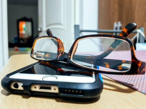 Eyeglasses with mobile phone on wooden table at night, close up