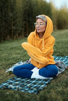 A cute girl with glasses in an orange hoodie is sitting in a park in nature expressing kind emotions