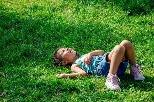 A little girl is smiling lying on the bright green grass. Portrait of a happy child on the grass in summer. The view from the top. Space for text