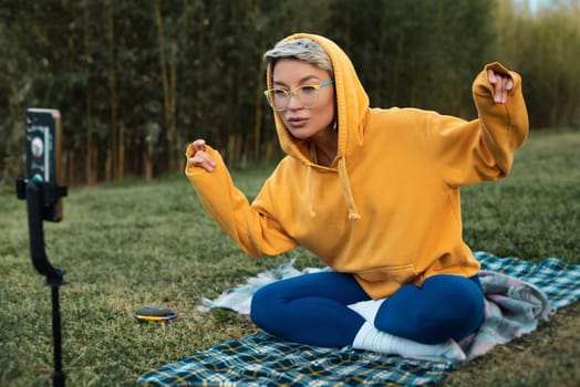 pretty blogger girl in glasses and an orange hoodie is sitting in a park in nature, streaming on her phone and emotionally chatting with subscribers, expressing kind emotions