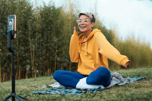 pretty blogger girl in glasses and an orange hoodie is sitting in a park in nature, streaming on her phone and emotionally chatting with subscribers, expressing kind emotions