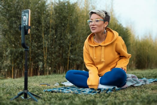 pretty blogger girl in glasses and an orange hoodie is sitting in a park in nature, streaming on her phone and emotionally chatting with subscribers, expressing kind emotions