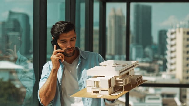 Smart caucasian engineer standing and holding house model while phone calling to manager. Professional manager inspect at architectural model while standing near window. Design concept. Tracery