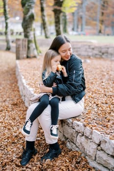 Mom hugs a little girl with an apple in her hands sitting on her lap on a low fence in the park. High quality photo