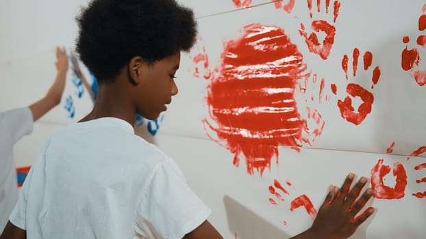 Side view of young children stamp hand with color and paint wall. Highschool student with mixed raced holding tray while paint room with red and blue in art lesson. Creative activity. Edification.