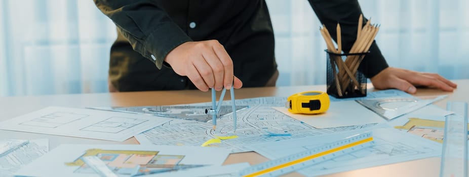 A portrait of architect using divider to measure blueprint. Architect designing house construction on a table at studio, with architectural equipment scattered around. Focus on hand. Delineation