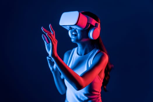 Smart female stand with surrounded by cyberpunk neon light wear VR headset connecting metaverse, futuristic cyberspace community technology. Woman using finger pointing virtual object. Hallucination.