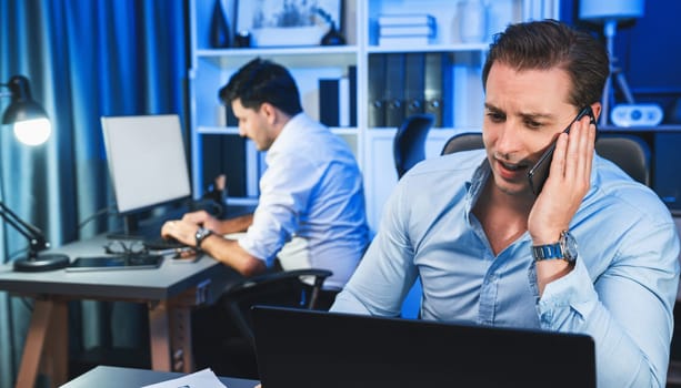 Serious businessman calling on phone with customer to create new project at night time, coworker writing document work sheet on desk. Concept of working on neon blue light modern office. Sellable.