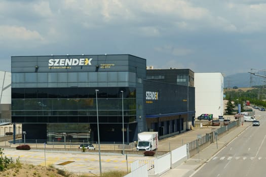 Barcelona, Spain - May 24, 2023:The modern headquarters building of Szendex Transport with a glass facade stands under a cloudy sky, accompanied by a parked delivery van and clear company signage.
