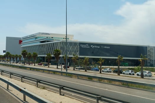 Barcelona, Spain - May 24, 2023: A sleek, contemporary office building stands under a clear sky, flanked by palm trees and parked cars, with a highway in the foreground.