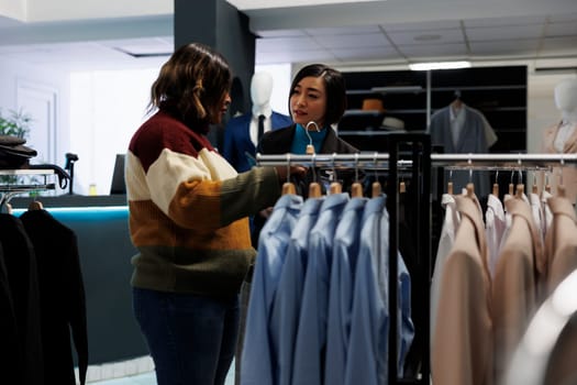 Clothing store woman asian assistant talking and consulting customer. Customer exploring rack with apparel, choosing formal outfit and getting help from boutique employee