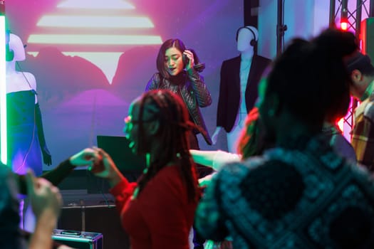 Woman dj performing on stage during disco party in nightclub. Asian musician holding headphones and mixing electronic music while crowd partying and dancing on dancefloor in club