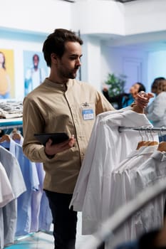 Clothing store assistant checking inventory with digital tablet and displaying apparel on rack. Fashion boutique manager hanging garment to attract customers in shopping center