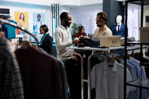 Smiling assistant holding hanger with stylish jacket, showcasing fashion trend to customer in clothing store. Boutique employee talking with client and helping to make apparel choice