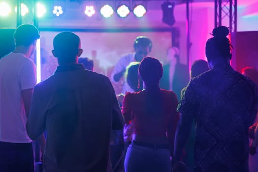 People partying and attending dj performance at nightclub discotheque. Young diverse men and women standing on crowded dark dancefloor while musician playing on stage in club