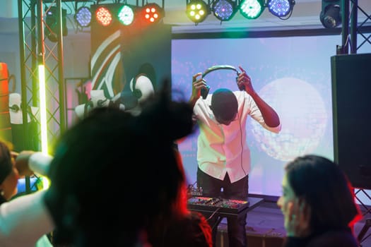 Dj wearing headphones on stage with spotlights during concert live performance in club. Musician putting on headset while mixing electronic music at disco party in nightclub