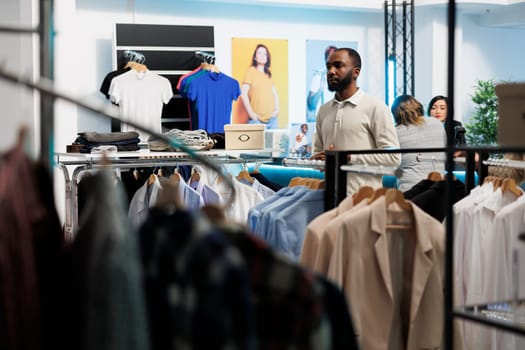African american man shopping for apparel and browsing rack while standing in clothing store. Department mall fashion boutique customer selecting casual outfit on hangers