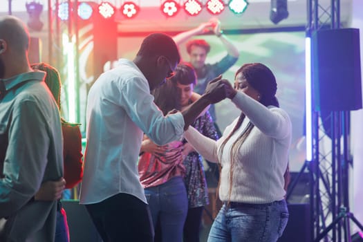 Young man and woman friends holding hands while dancing at nightclub party. African american couple clubbing and moving on dancefloor while attending discotheque gathering event