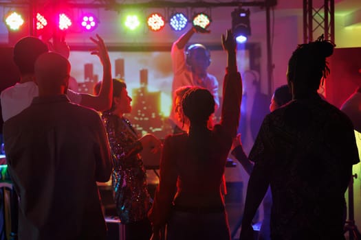 Clubbers partying and dancing while attending dj performance in nightclub. Young people crowd standing with raised hands on dancefloor illuminated with spotlights at discotheque