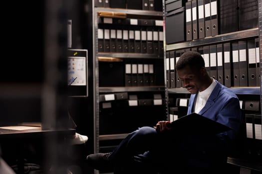African american manager organizing corporate files, analyzing bureaucracy record in arhive room. Businessman wearing formal suit checking accountancy documents in office depository