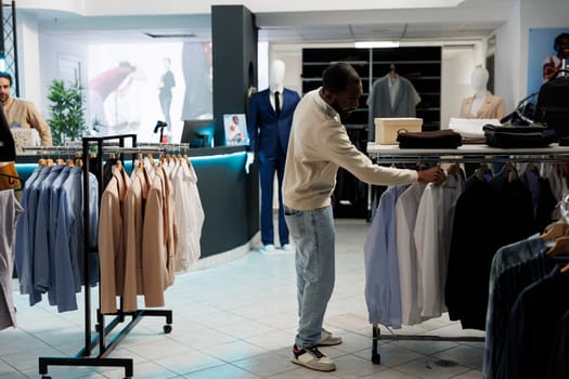 African american man searching shirt size while choosing formal wear outfit in clothing store. Boutique customer browsing rack with hanging apparel in shopping mall outlet