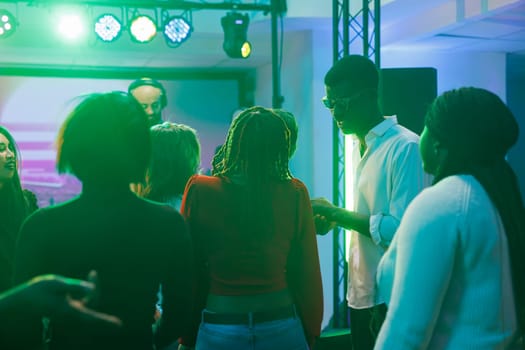 People partying and having fun at social gathering while attending dj concert in nightclub. Diverse crowd relaxing on dancefloor while musician performing on stage in dark club