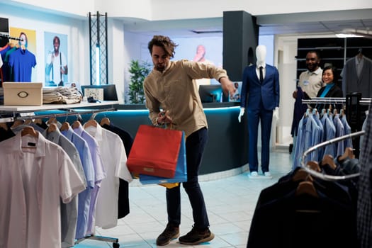 Happy young caucasian man dancing with paper packages in shopping center after buying fashionable apparel. Satisfied joyful customer carrying bags with purchased trendy outfit