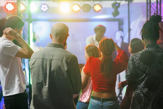 Young people standing on dancefloor while attending electronic music concert in nightclub. Diverse men and women crowd dancing while dj performing on stage with colorful spotlights