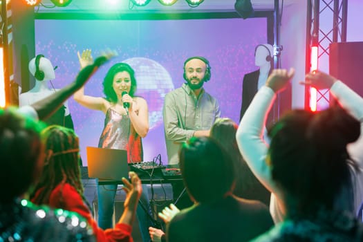 Young man and woman band performing on stage in nightclub at techno music festival. People partying on dancefloor while enjoying singer and dj playing at live concert in club